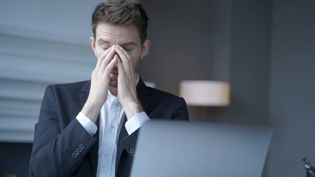 Overworked male entrepreneur sits at home office, suffering from heavy headache and fatigue