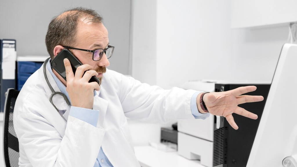 Male doctor pointing with finger on desktop computer while talking on the phone, discussing treatment with colleague.